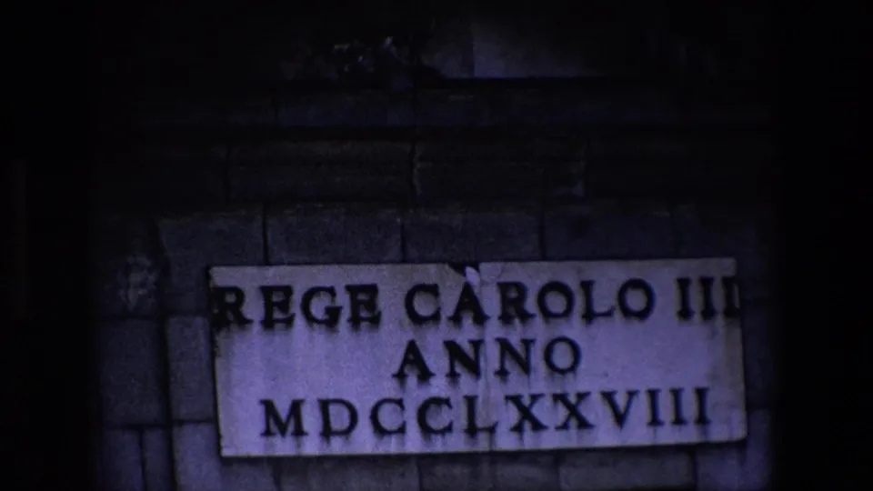 Madrid 1967 home-movie frame showing the plaque in lower exposure, with bold Roman-style text and numerals partly surrounded by darkened stonework and film borders.