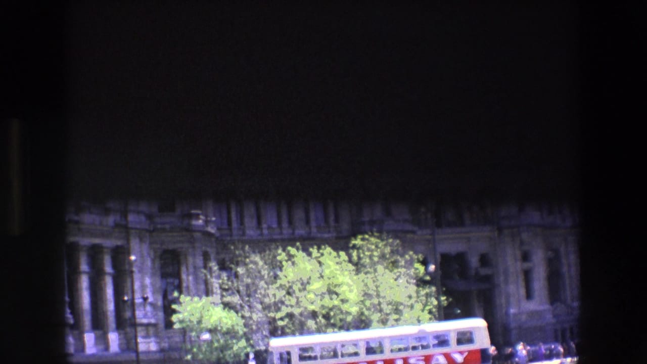 Underexposed stone building facade behind trees with a bus crossing the lower frame.