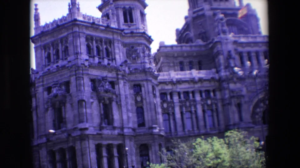 1967 Madrid home-movie frame with a tilted view of an ornate stone civic building, tower-like corner detail, arched entry area, and trees at street level.