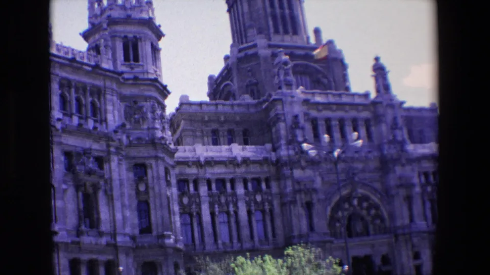 1967 Madrid frame of a monumental stone building with vertical window bays, a large arched entrance, a tower, a small Spanish flag, and trees in the foreground.