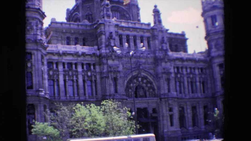 1967 Madrid 8mm frame centered on the ornate stone facade, large arched doorway, rows of windows, foreground trees, and part of a street lamp.