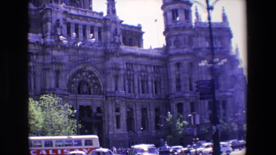 1967 Madrid frame showing the monumental stone entrance and tower with plaza trees, a bus, cars, street lamps, and directional signs in the foreground.