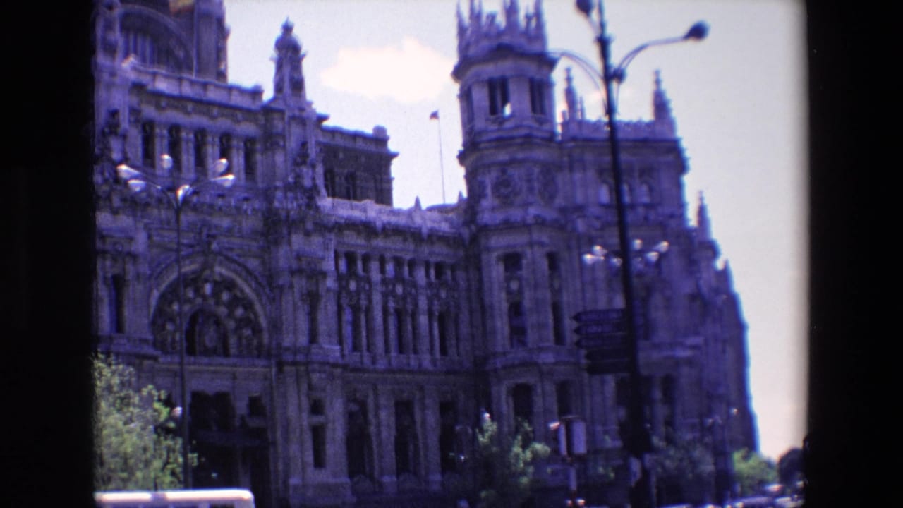 Ornate stone landmark facade with towers, streetlamps, trees, and a bright sky.