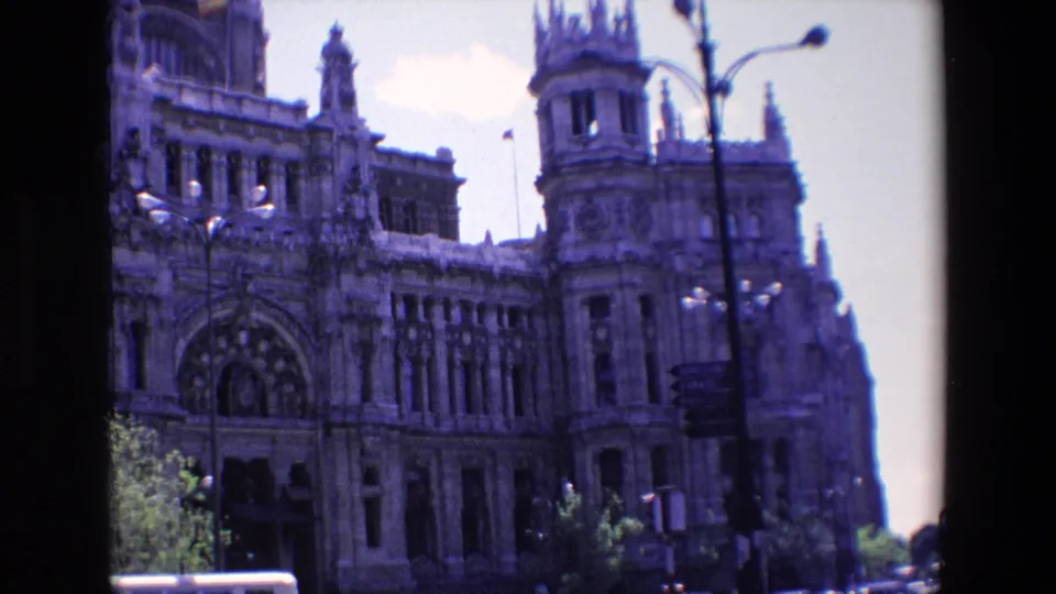 1967 Madrid home-movie frame showing the Palacio de Cibeles facade from street level, with decorative towers, streetlamps, trees, and traffic cropped along the bottom edge.