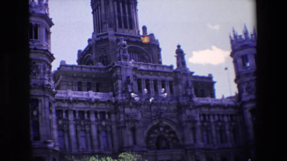 Tilted 1967 Madrid home-movie frame showing Palacio de Cibeles with its clock tower, Spanish flag, arched entrance, decorative roofline, and street-level foreground.