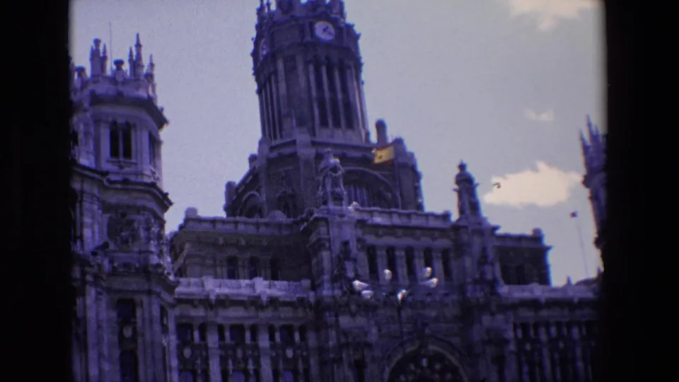 1967 Madrid frame centering the Palacio de Cibeles clock tower and upper facade, with the Spanish flag, streetlamps, and open sky in a handheld 8mm view.