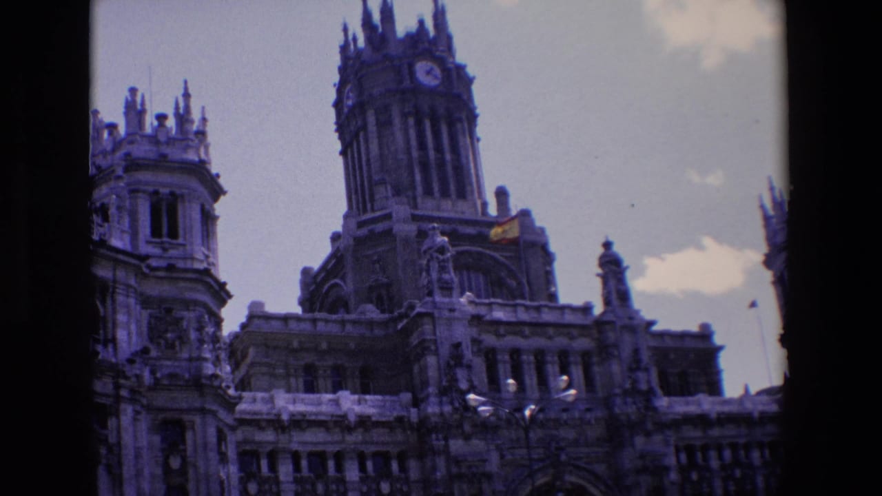 Ornate building facade with a clock tower, spires, and a Spanish flag against the sky.