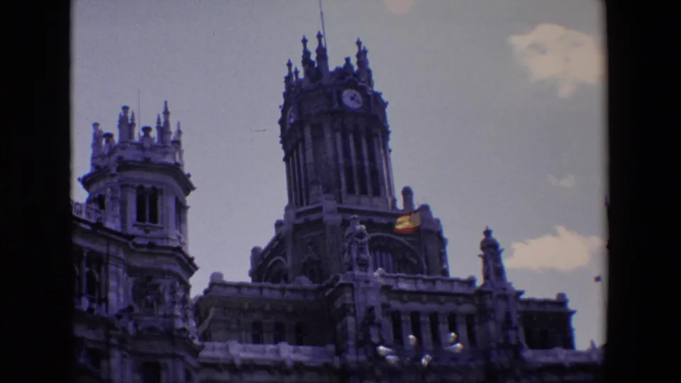 1967 Madrid frame with the ornate tower centered, a clock face near the top, flanking spires, and a Spanish flag catching light on the building.