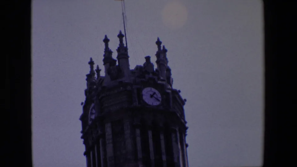 1967 Madrid frame isolating the upper clock tower, clustered spires, and flagpole against a plain sky in soft, dark 8mm exposure.