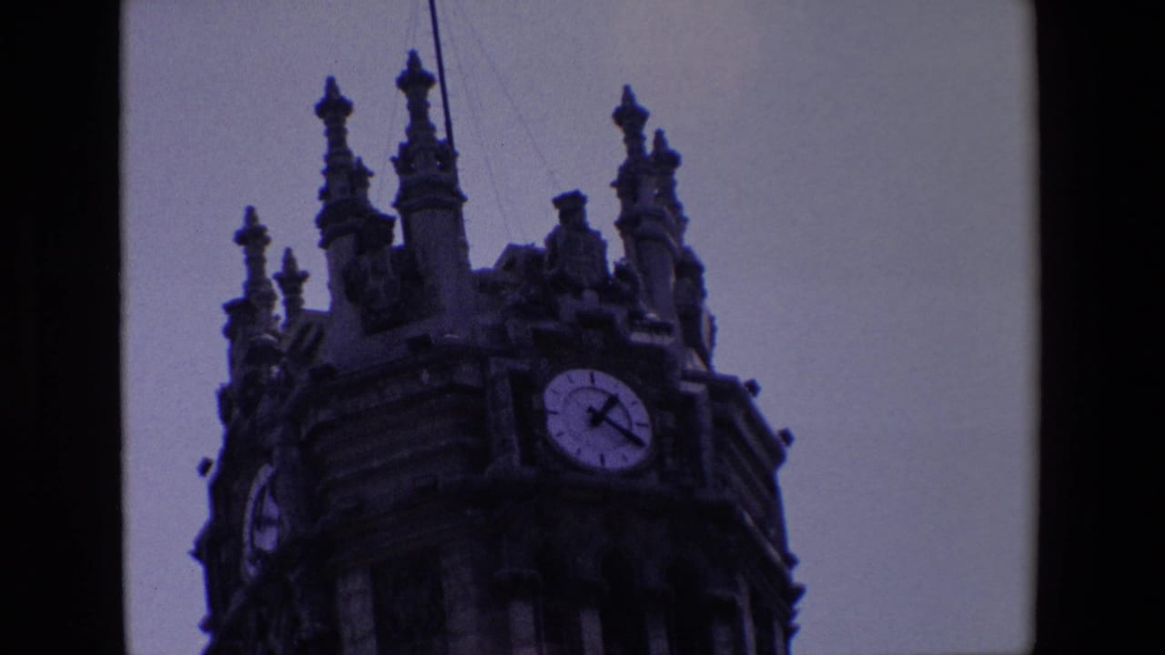 1967 Madrid 8mm frame showing a low-angle visitor view of an ornate stone clock tower with a visible round clock face and dark film edges.