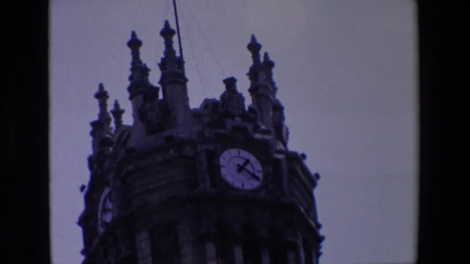 1967 Madrid 8mm frame showing a low-angle visitor view of an ornate stone clock tower with a visible round clock face and dark film edges.