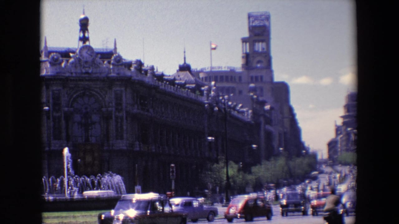 Traffic moves past a fountain and long ornate building on a sunlit city street.