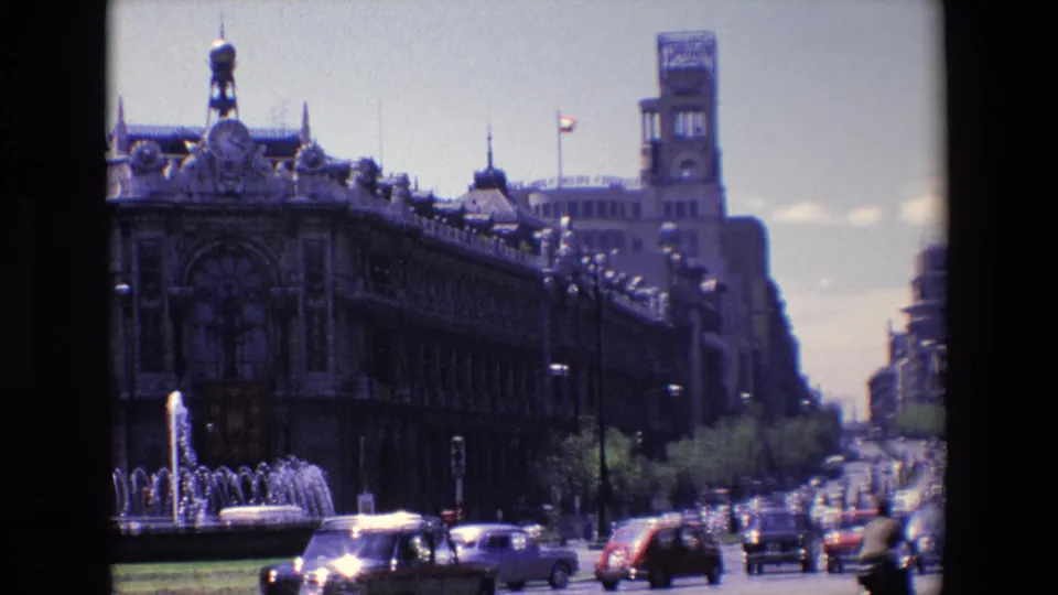 1967 Madrid traffic passes a foreground fountain and long ornate building with a tower, with more city buildings receding down the avenue.