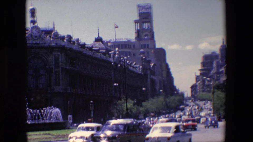 1967 Madrid avenue view with classic cars, fountain spray, a dark ornate facade, trees, and distant buildings framed by 8mm film edges.