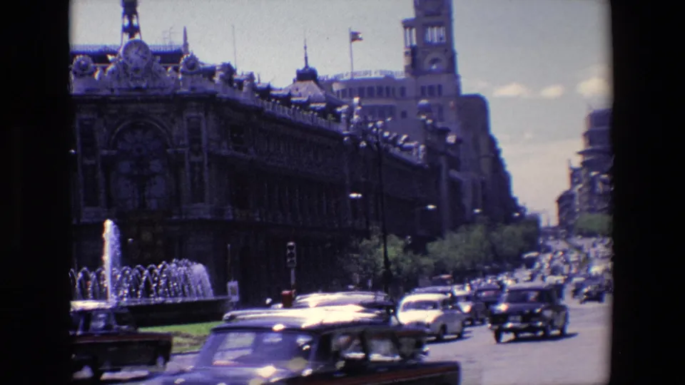 1967 Madrid street traffic fills the foreground as cars pass a water fountain and ornate building with a tower along a busy urban avenue.