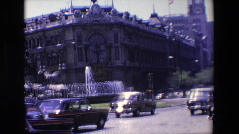 In 1967 Madrid, dark cars move past a public fountain with sculpted figures and a large ornate building in the background.