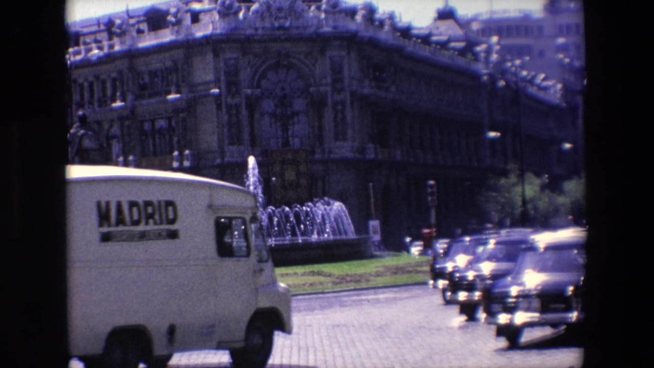 A white van marked Madrid passes traffic near a fountain and large building.