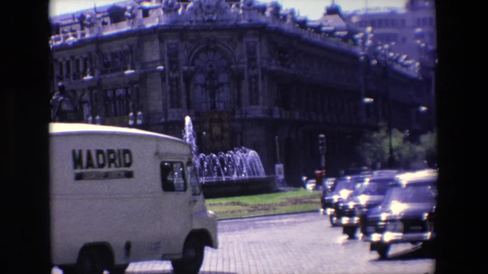 A white delivery van marked Madrid drives through 1967 Madrid street traffic beside a fountain and a large ornate building.