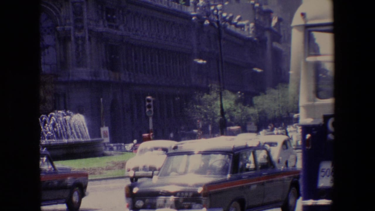 Classic cars and a bus pass a fountain and large historic building on a busy city street.