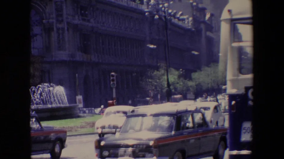 1967 Madrid street traffic with classic sedans, a bus, a fountain, traffic lights, trees, and an ornate building facade in the background.