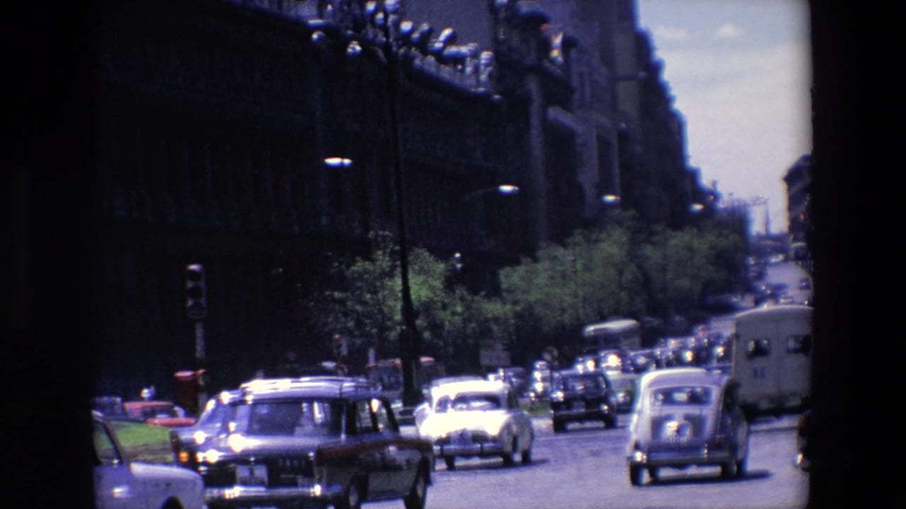 Cars and buses move along a broad street beside a long ornate building facade.