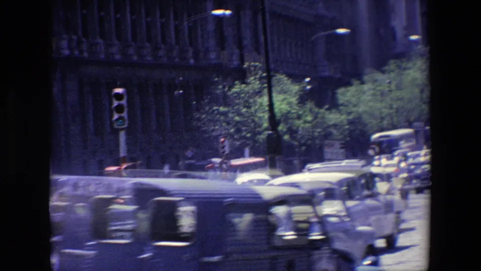 1967 Madrid roadway with motion-blurred classic vehicles, traffic signals, trees, street lamps, and a tall masonry facade behind the traffic.