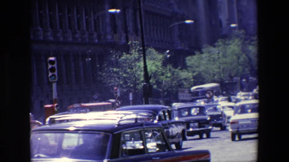 1967 Madrid street traffic with a dark station wagon in the foreground, classic cars, buses, traffic lights, trees, and a large historic facade.
