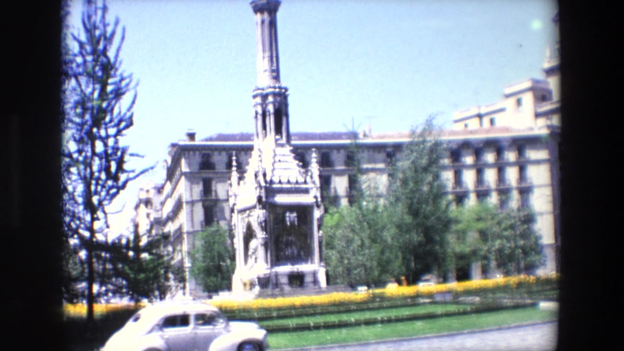 Ornate stone monument in a landscaped square with a white car passing in front.