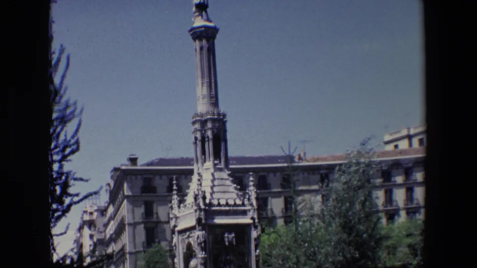 1967 Madrid home-movie frame with a tall column and ornate monument base, bordered by trees, lawns, flower beds, and urban buildings.