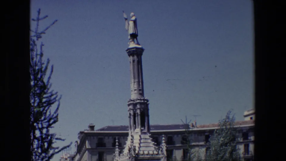 1967 Madrid frame emphasizing the tall monument column and statue, with tree branches, building rooflines, and blue sky around the public square.