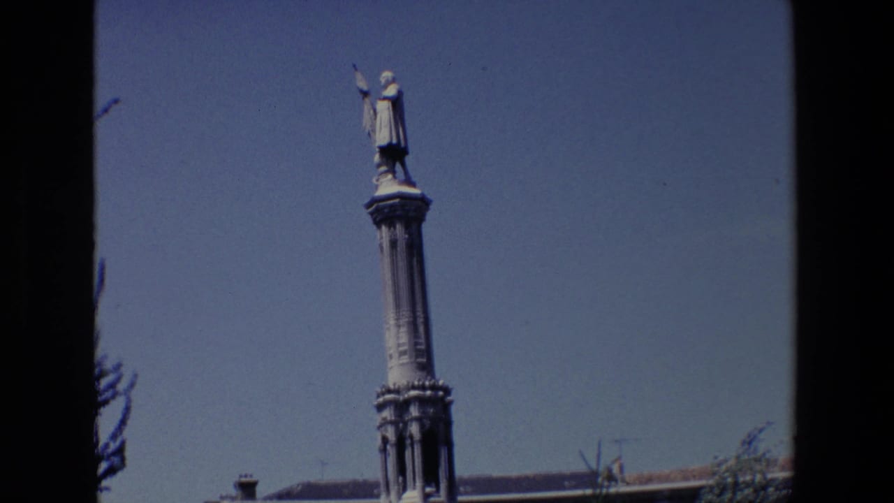 1967 Madrid 8mm frame showing a tall column topped by a robed male statue holding a flag, with blue sky and a low rooftop edge below.