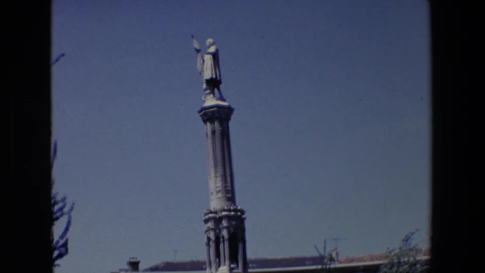 1967 Madrid 8mm frame showing a tall column topped by a robed male statue holding a flag, with blue sky and a low rooftop edge below.