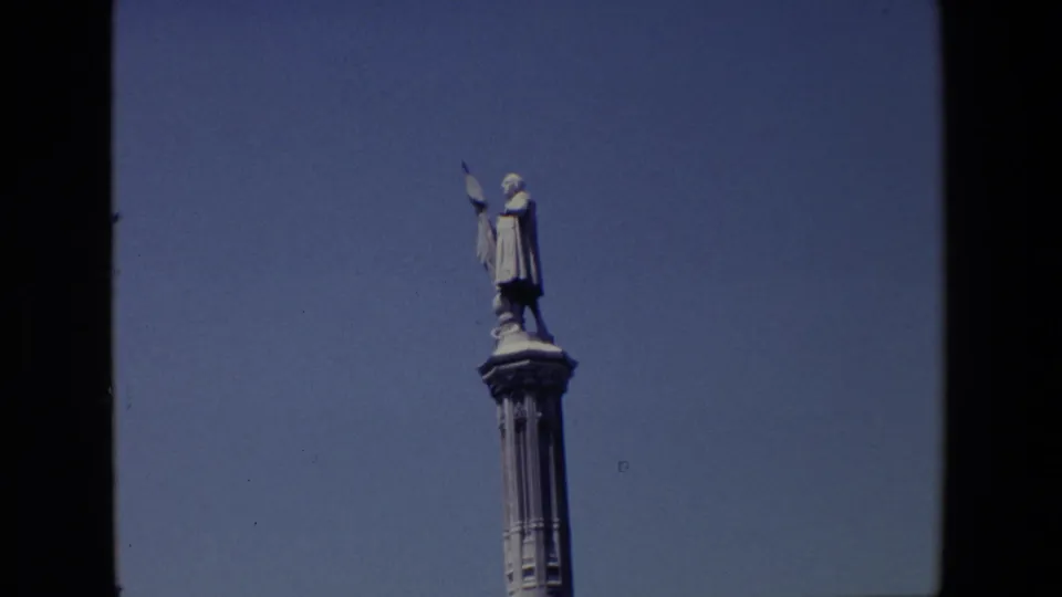 1967 Madrid 8mm frame with the robed statue and flag seen from below on a tall decorative pedestal, isolated against blue sky.