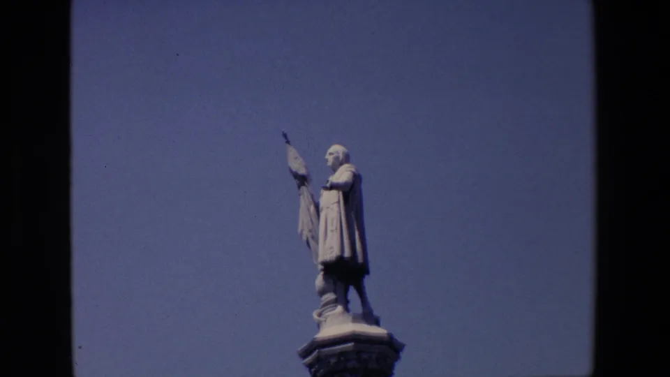 1967 Madrid 8mm close view of the robed figure holding a flag while standing on the top of the monument pedestal against a clear sky.