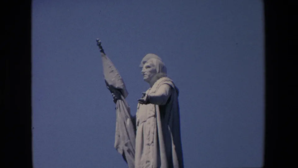 1967 Madrid 8mm close-up showing the upper body of a robed male statue with one arm extended and a flag or banner held upright against the sky.
