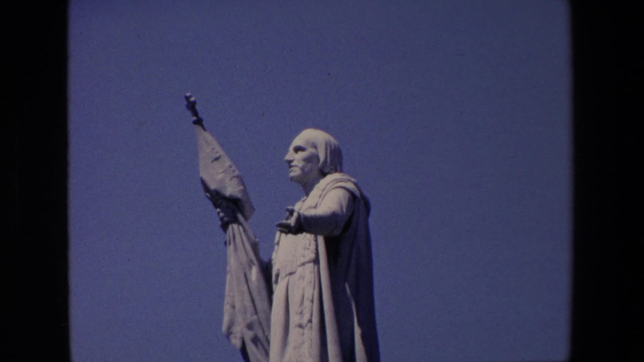 1967 Madrid home-movie frame showing a large robed stone religious statue in profile, holding a raised banner against a clear blue sky.