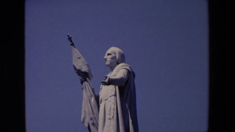 1967 Madrid home-movie frame showing a large robed stone religious statue in profile, holding a raised banner against a clear blue sky.