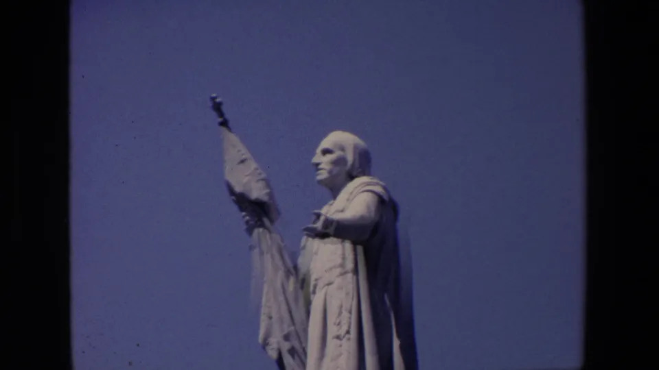 1967 Madrid 8mm frame of a large stone saint-like figure in profile, draped in robes and holding an upright banner against open sky.