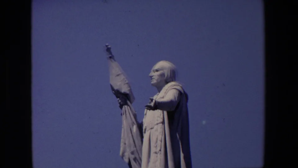 1967 Madrid home-movie still showing the robed stone figure from below, with the raised banner and clear sky emphasizing the monument's height.