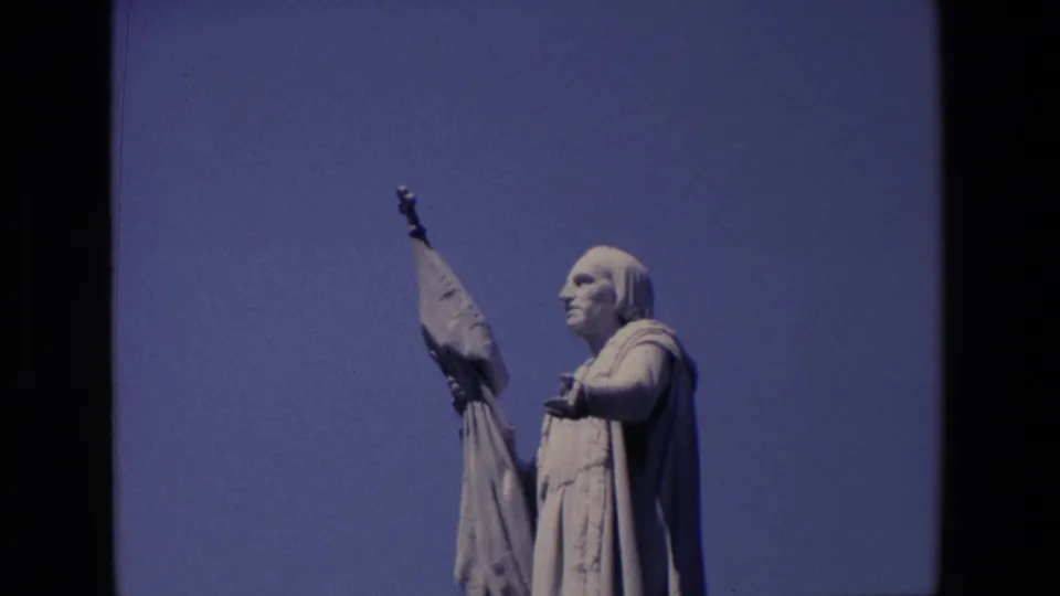 1967 Madrid 8mm frame of the stone religious statue in profile, with a robe, raised banner, and uncluttered blue sky background.