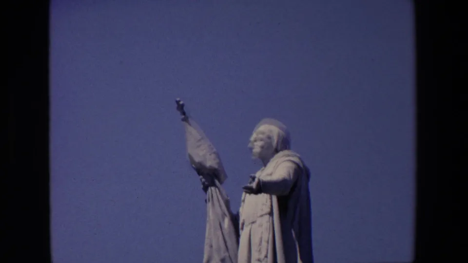 1967 Madrid 8mm frame showing a pale stone statue of a robed figure holding a flag against a blue sky, with dark film borders visible.