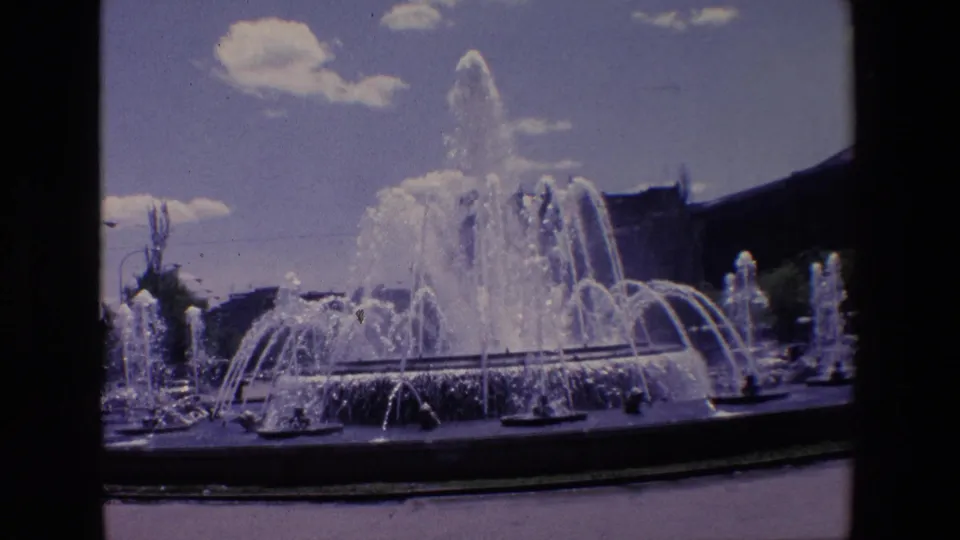 1967 Madrid 8mm frame showing a public fountain with multiple water jets, a central spray, low buildings, and small clouds in bright daylight.
