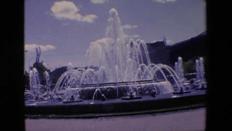 1967 Madrid 8mm frame of a broad fountain basin with clustered jets, a high central plume, partly cloudy sky, and surrounding urban structures.