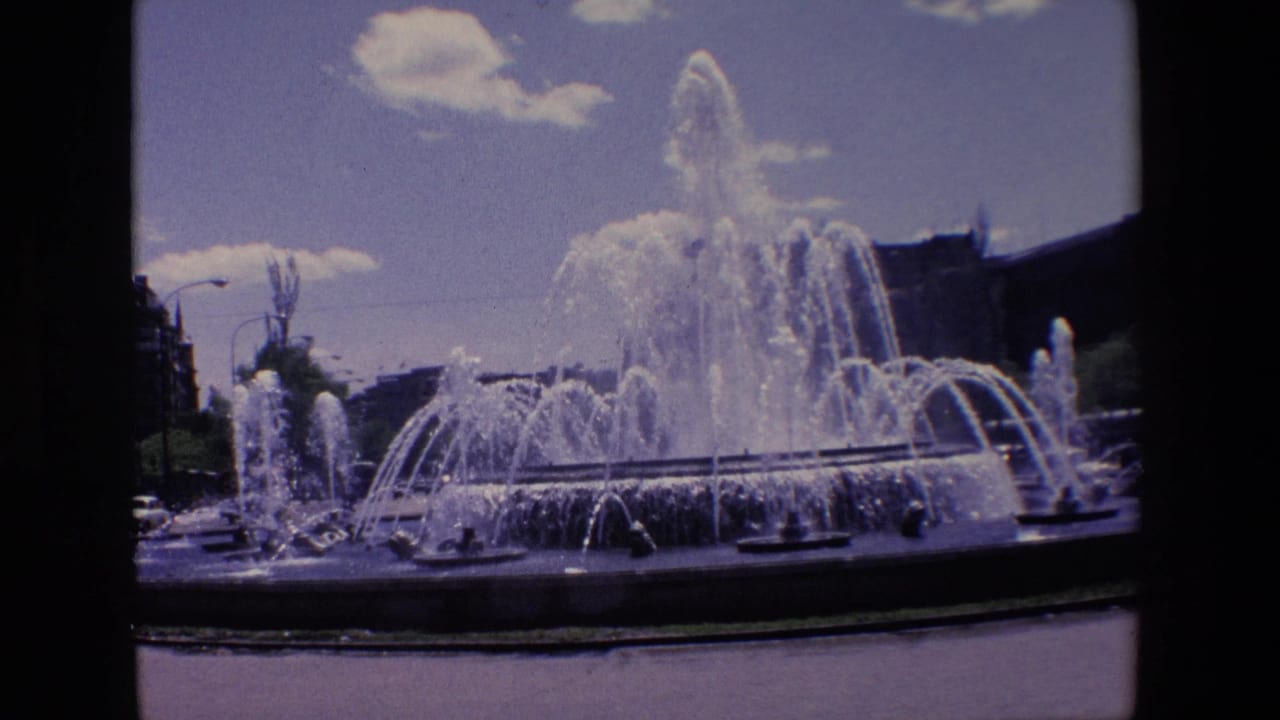 1967 Madrid home-movie frame showing a large public fountain with multiple water jets, background buildings, streetlights, and visible 8mm frame edges.