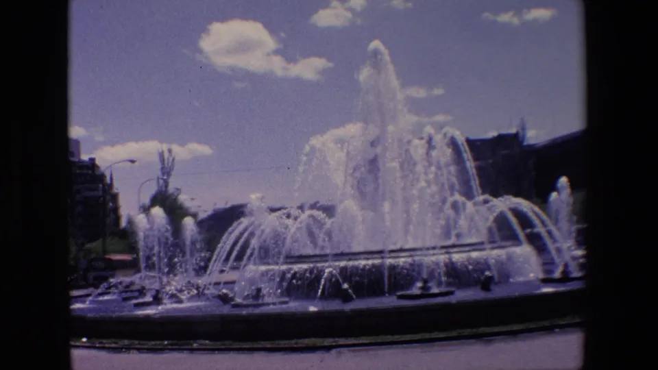 1967 Madrid frame of a large urban fountain, with a tall central plume, lower arcing jets, clouds overhead, and building silhouettes behind it.
