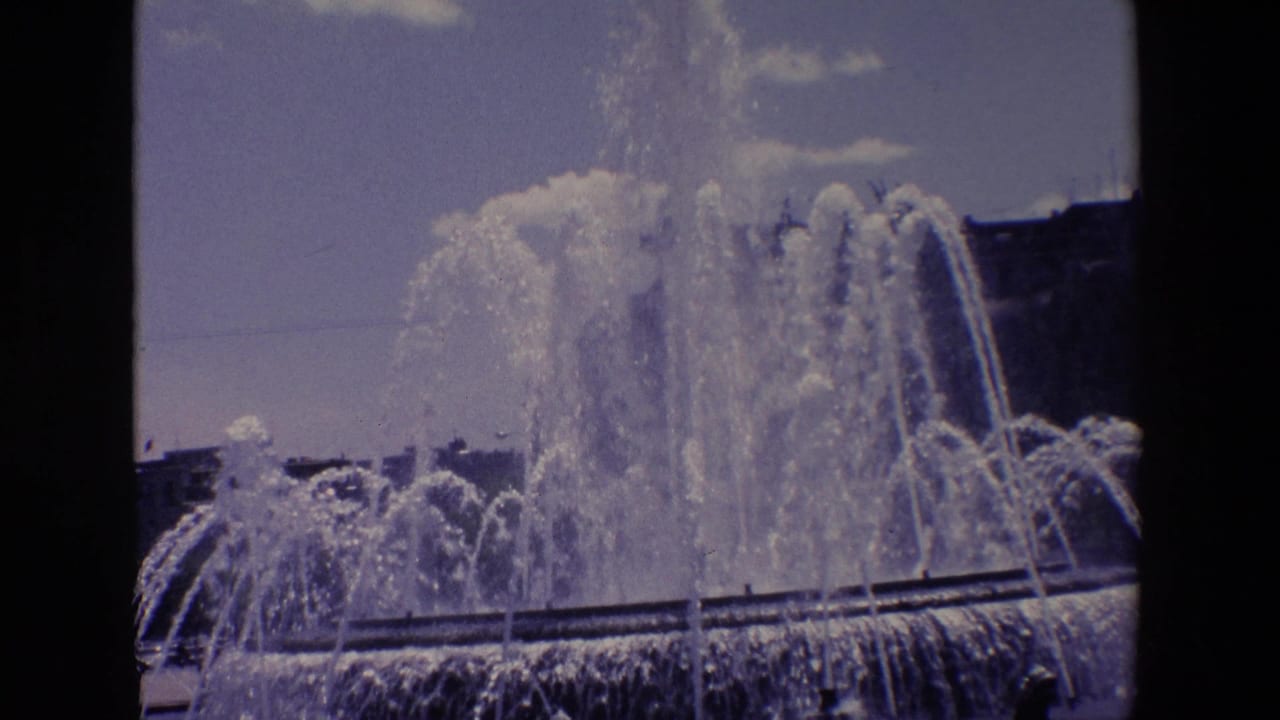 1967 Madrid 8mm frame showing a large public fountain with high water jets, a basin edge, sky, and urban buildings in the background.