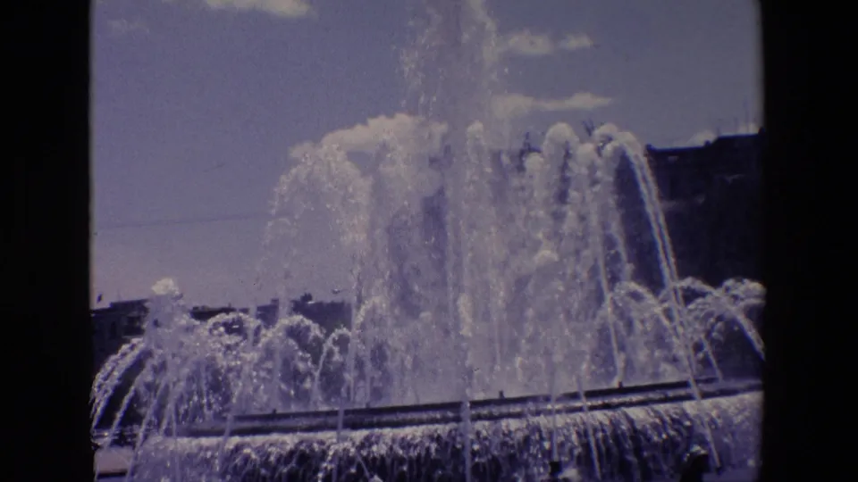 1967 Madrid 8mm frame showing a large public fountain with high water jets, a basin edge, sky, and urban buildings in the background.