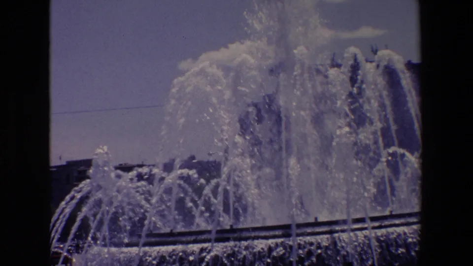 1967 Madrid 8mm frame of dense fountain spray and curved jets crossing a public basin, with city structures visible beyond the water.