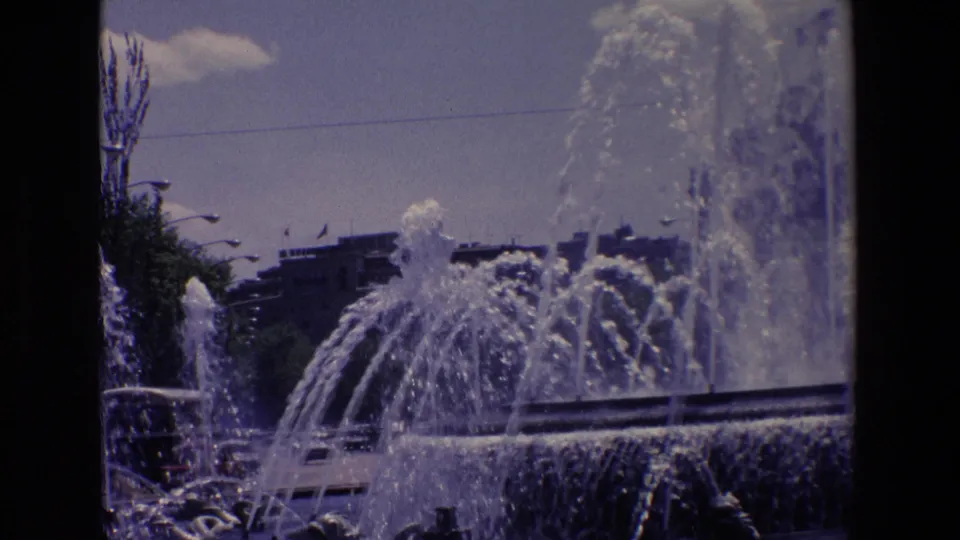 1967 Madrid 8mm frame showing fountain spray beside streetlights, trees, cars, and apartment-like buildings beyond the plaza.