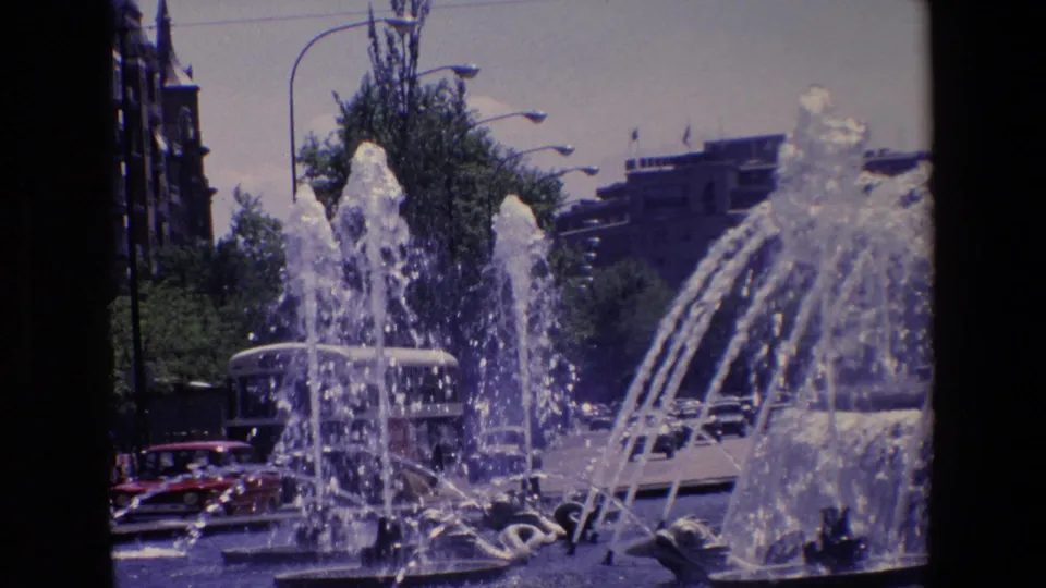 1967 Madrid 8mm frame with foreground fountain jets, visible cars near the basin, trees, streetlights, and urban buildings in the background.